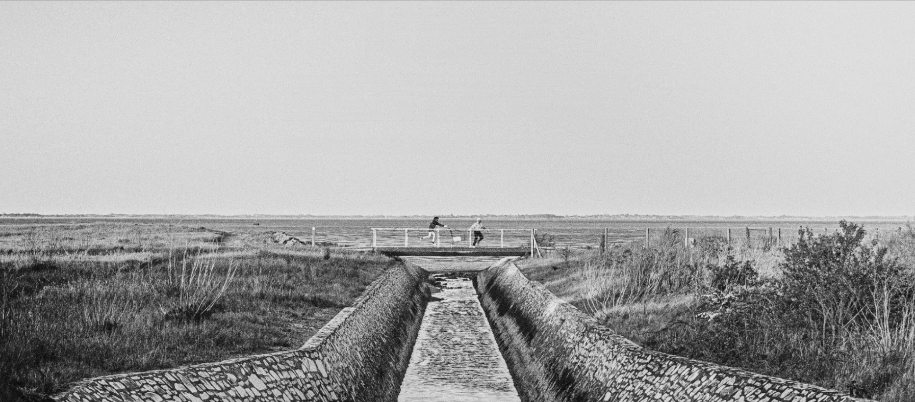 Deux silhouettes traversent une passerelle au-dessus d’un canal, dans un paysage de marais en noir et blanc